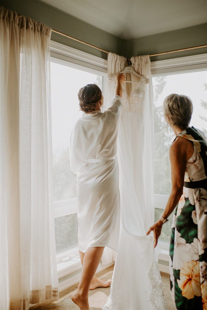 Bride reaching for wedding dress hanging by the window while mother helps with the veil